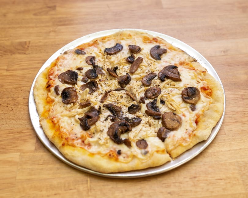 A whole mushroom and chicken pizza on a metal pan, resting on a wooden table.