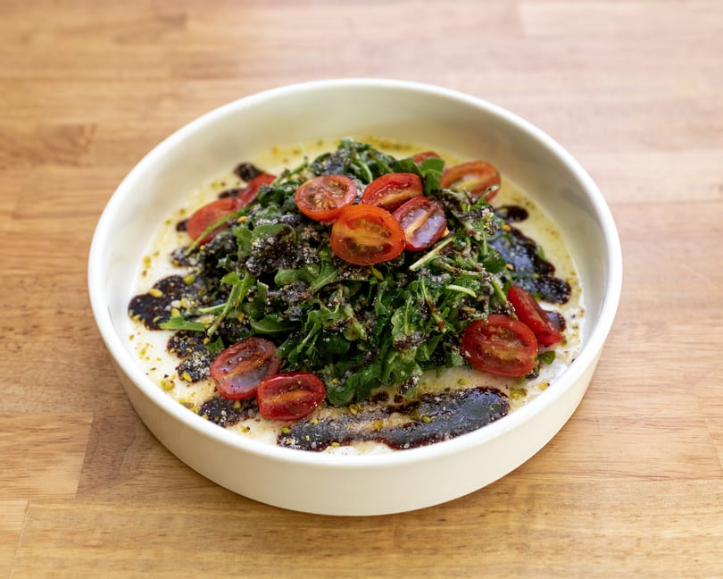Arugula salad with halved cherry tomatoes, dark dressing, and cheese in a white bowl.