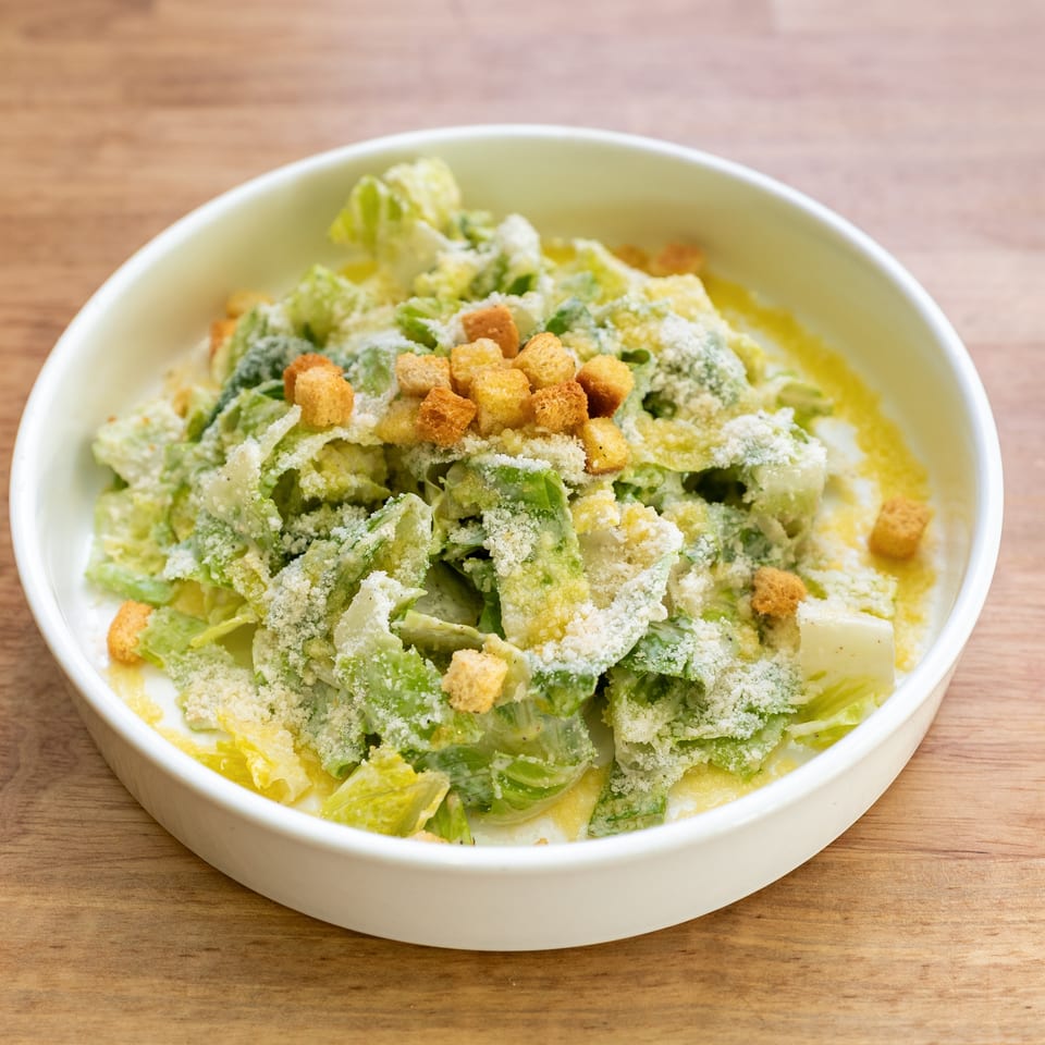 A white bowl filled with Caesar salad, topped with croutons and grated Parmesan cheese.
