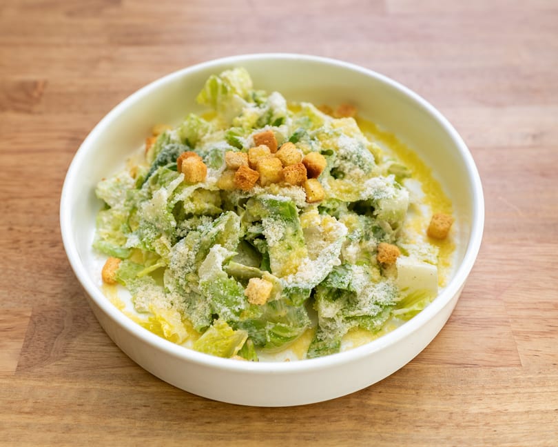 A white bowl filled with Caesar salad, topped with croutons and grated Parmesan cheese.