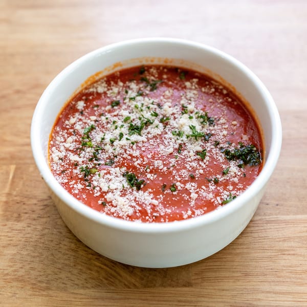 A white bowl of red tomato soup topped with grated cheese and fresh herbs on a wooden table.
