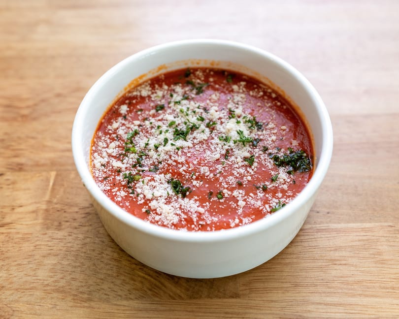 A white bowl of red tomato soup topped with grated cheese and fresh herbs on a wooden table.