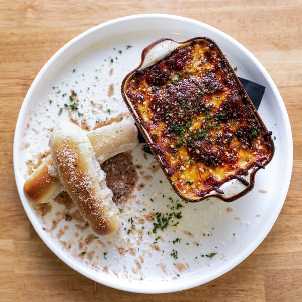 Baked pasta in a small dish with two breadsticks on a white plate, on a wooden table.