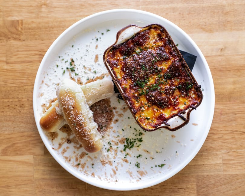 Baked pasta in a small dish with two breadsticks on a white plate, on a wooden table.