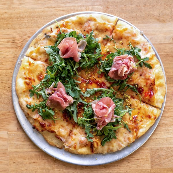 A whole pizza topped with arugula, prosciutto, and pistachios on a metal tray.