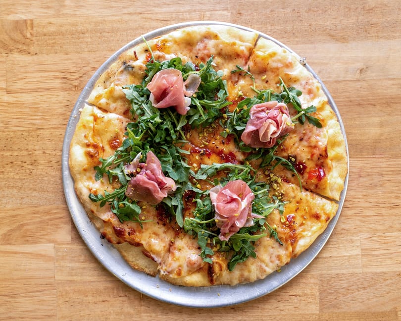 A whole pizza topped with arugula, prosciutto, and pistachios on a metal tray.