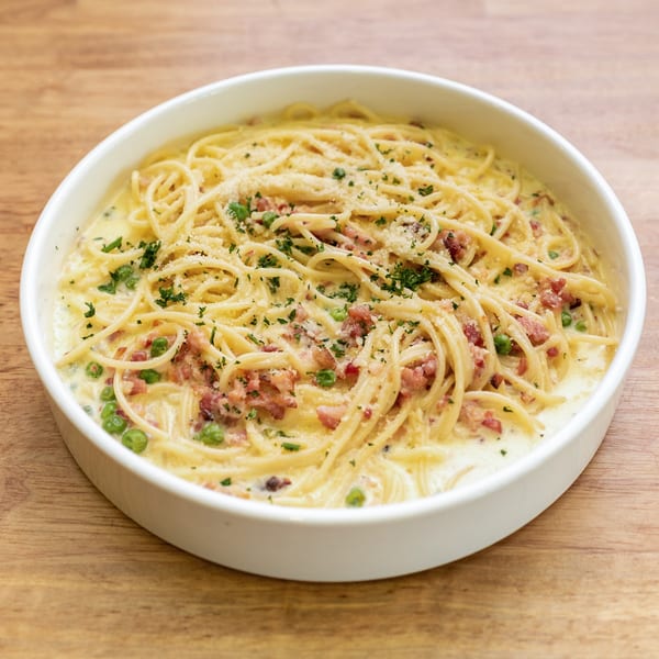 Creamy spaghetti carbonara with bacon, peas, and parsley in a white bowl on a wooden table.