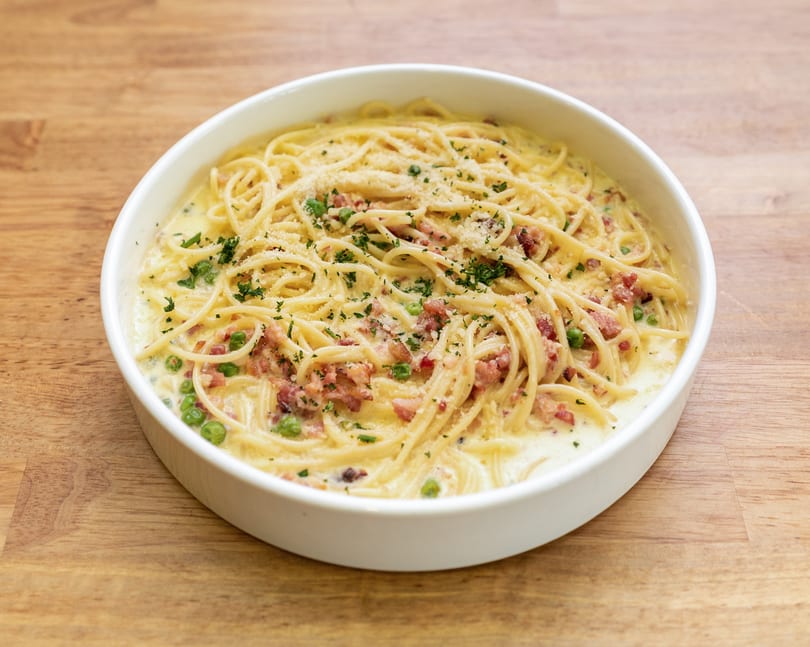 Creamy spaghetti carbonara with bacon, peas, and parsley in a white bowl on a wooden table.