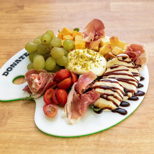 Charcuterie board with grapes, cheeses, prosciutto, tomatoes, burrata, and balsamic glaze on a wooden table.