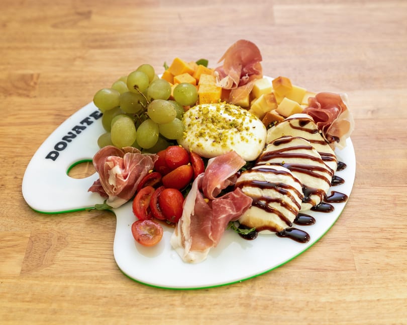 Charcuterie board with grapes, cheeses, prosciutto, tomatoes, burrata, and balsamic glaze on a wooden table.