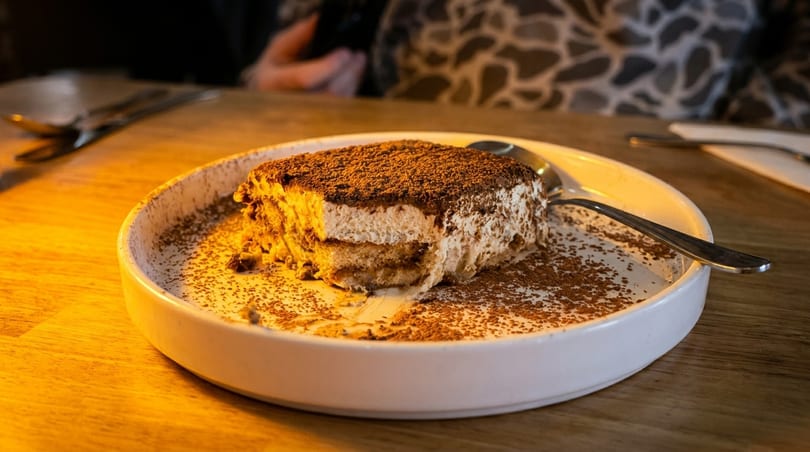 A partially eaten tiramisu dessert, dusted with cocoa powder, on a white plate with a spoon.