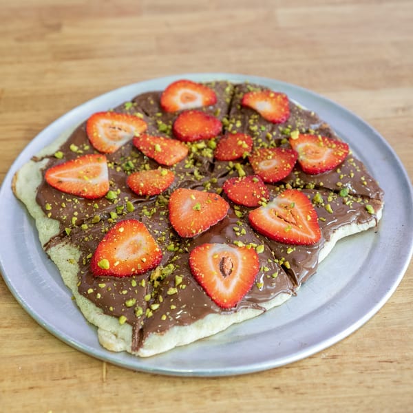 Dessert pizza with chocolate spread, sliced strawberries, and chopped pistachios on a metal plate.