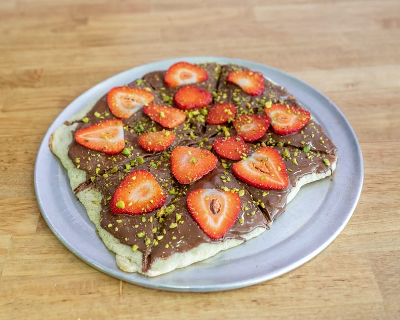 Dessert pizza with chocolate spread, sliced strawberries, and chopped pistachios on a metal plate.