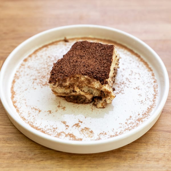 A slice of tiramisu dessert, topped with cocoa powder, on a white plate with sprinkled cinnamon.