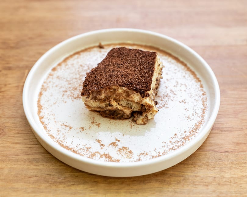 A slice of tiramisu dessert, topped with cocoa powder, on a white plate with sprinkled cinnamon.