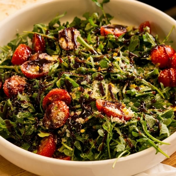 Arugula salad with cherry tomatoes, parmesan cheese, and balsamic glaze in a white bowl.