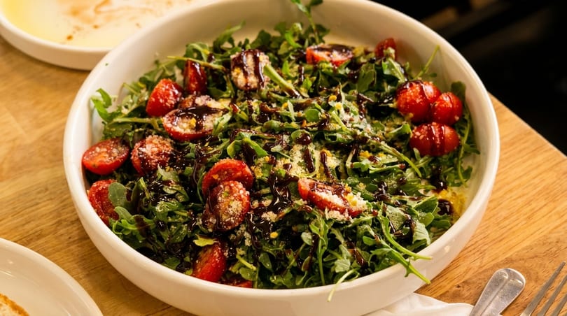 Arugula salad with cherry tomatoes, parmesan cheese, and balsamic glaze in a white bowl.