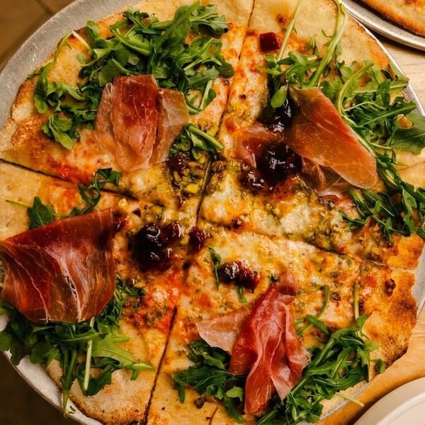 Overhead view of a pizza topped with prosciutto, arugula, and a dark jam, sliced on a metal tray.