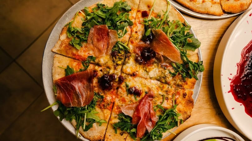 Overhead view of a pizza topped with prosciutto, arugula, and a dark jam, sliced on a metal tray.