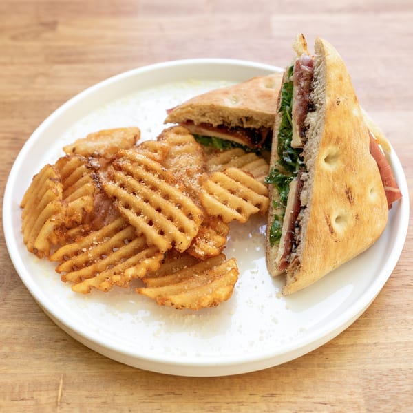 Focaccia sandwich with prosciutto and arugula, served with waffle fries on a white plate.
