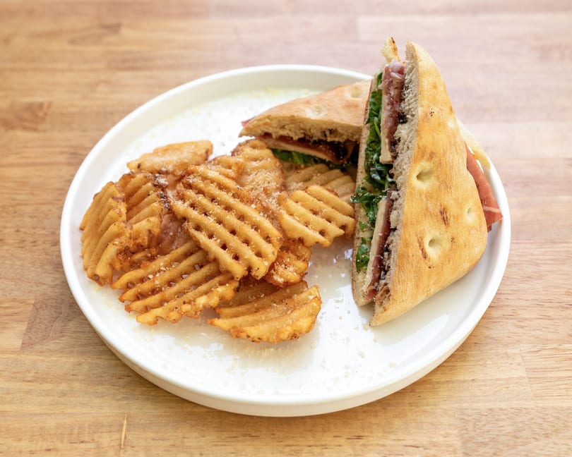 Focaccia sandwich with prosciutto and arugula, served with waffle fries on a white plate.
