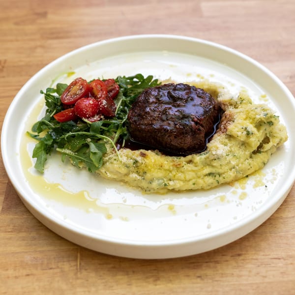 A gourmet dish featuring a steak medallion on mashed potatoes, with arugula and cherry tomatoes.