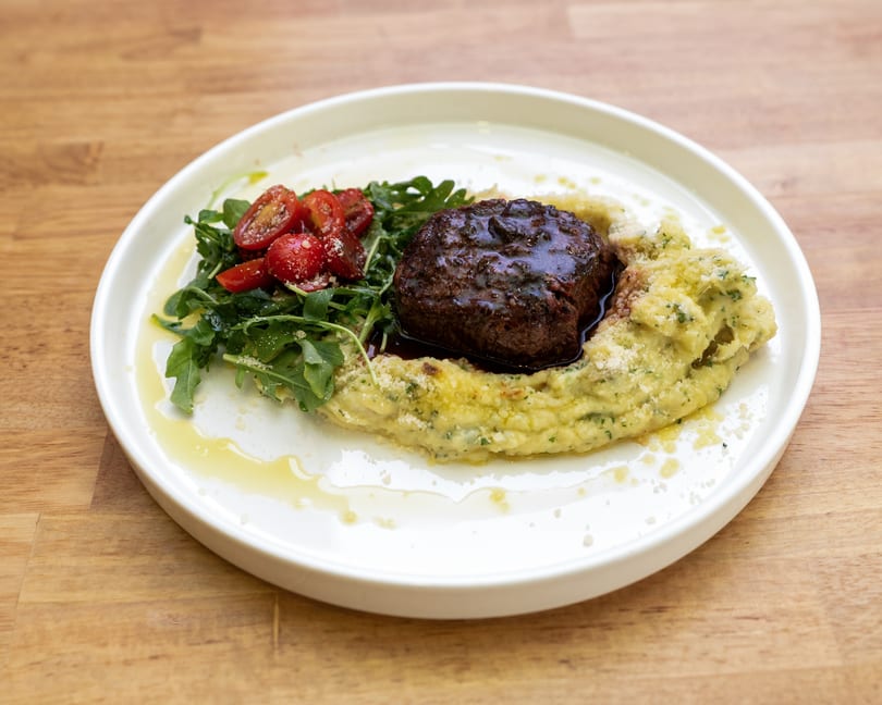 A gourmet dish featuring a steak medallion on mashed potatoes, with arugula and cherry tomatoes.