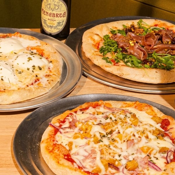 Three pizzas on metal trays with two bottles of Menabrea beer on a wooden table.