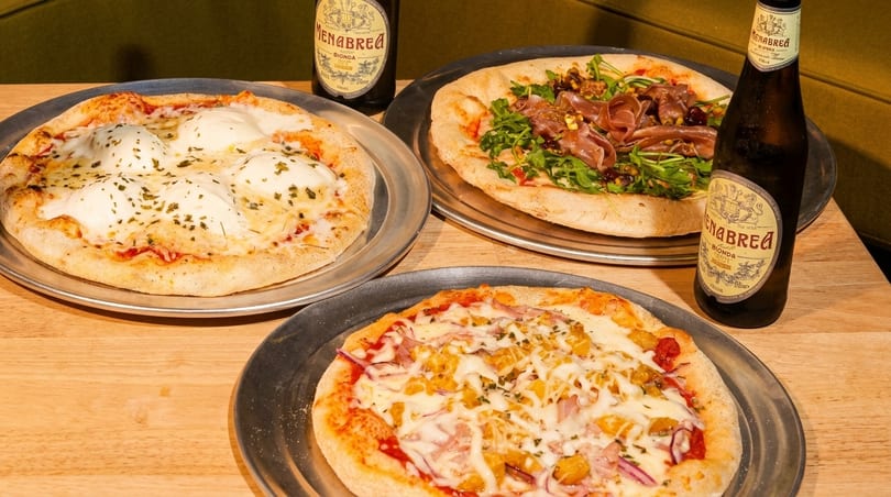 Three pizzas on metal trays with two bottles of Menabrea beer on a wooden table.