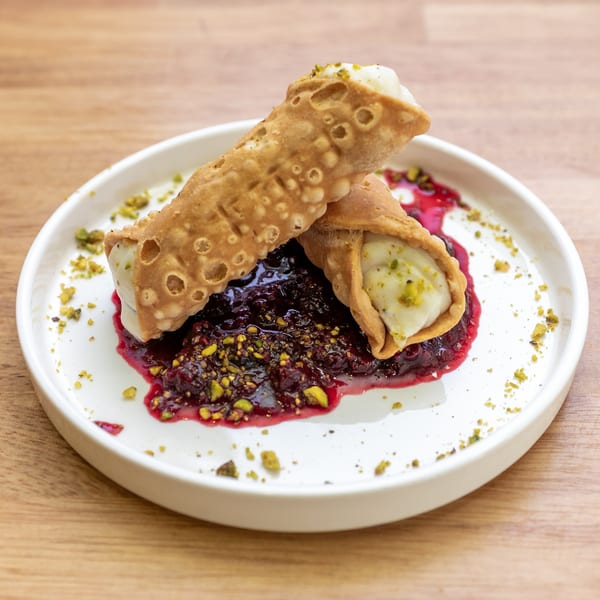 Two cannoli filled with cream and pistachios, served on a white plate with berry compote.