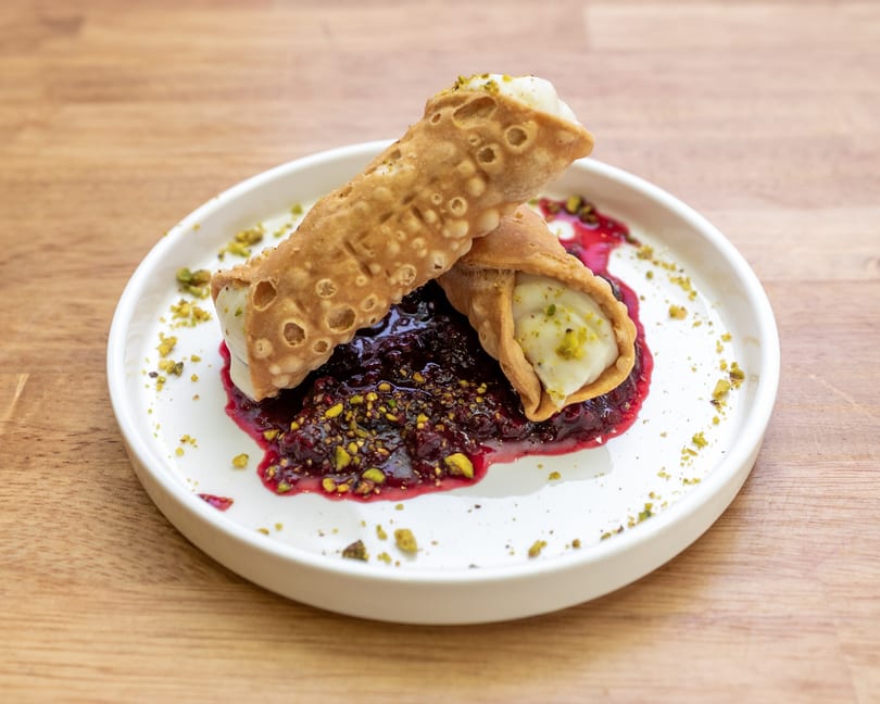 Two cannoli filled with cream and pistachios, served on a white plate with berry compote.